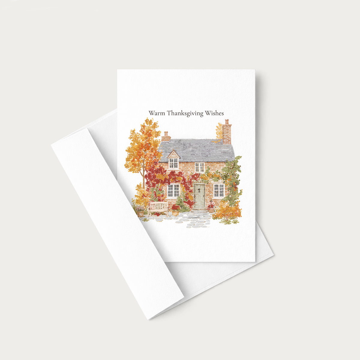 A white Thanksgiving greeting card with a watercolor illustration of a cottage surrounded by autumn foliage, shown with a matching white envelope on a light gray background.