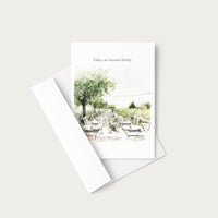 A white greeting card with a watercolor illustration of an outdoor dining scene and the printed phrase 'Today, we become family,' shown with a white envelope on a light gray background.