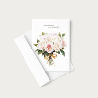 A white greeting card with a watercolor bouquet of pale pink and white roses, green leaves, and a gold ribbon, with the printed message 'Dearest Mother, on my wedding day.' It is shown with a white envelope on a light warm gray background.