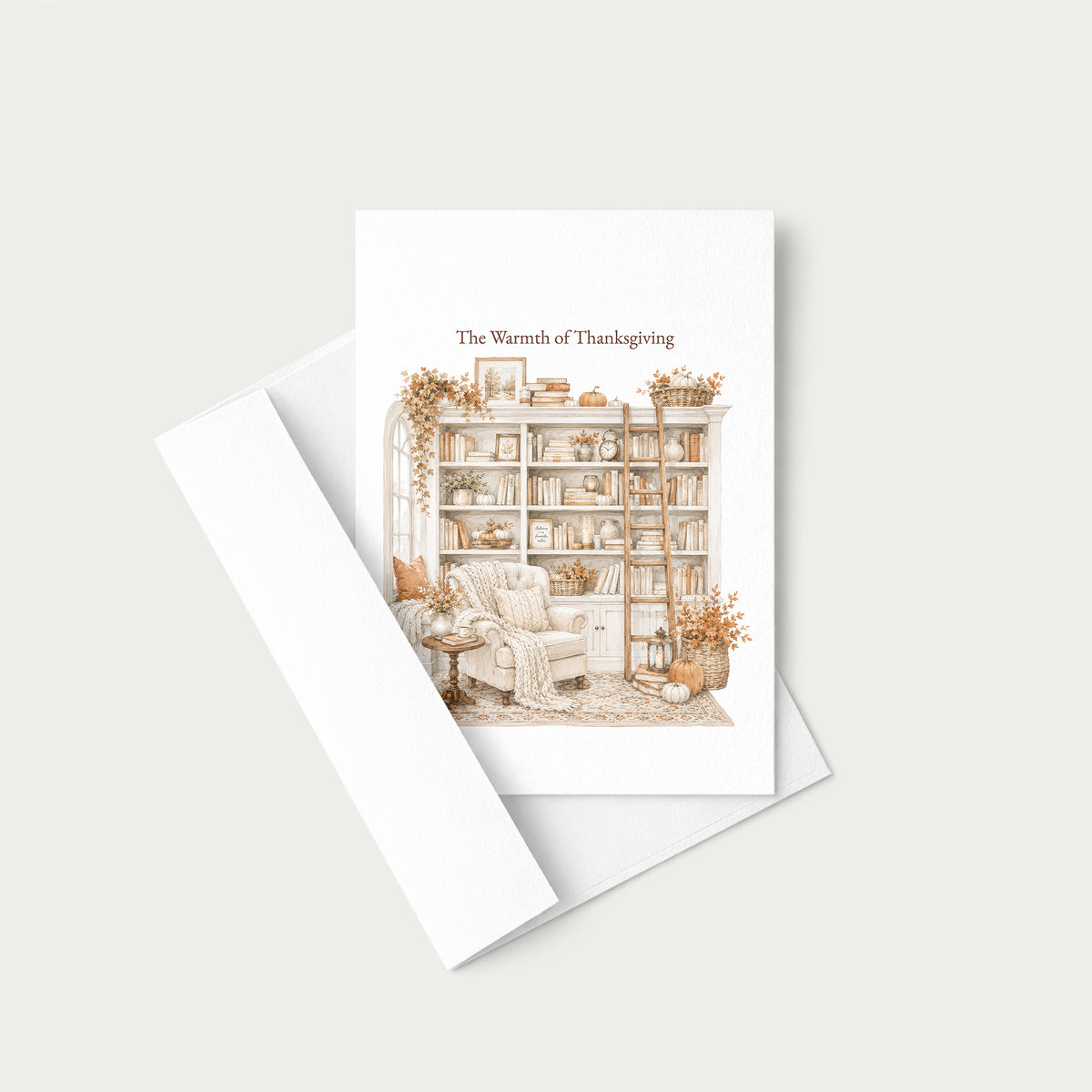A white greeting card with the text 'The Warmth of Thanksgiving' and a delicate illustration of a cozy reading nook with a bookshelf, armchair, and autumn decor, shown with a white envelope on a light beige background.