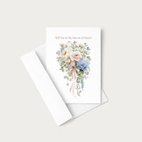 A white wedding greeting card with a watercolor bouquet illustration and the printed phrase 'Will You Be My Mateon of Honor?' in dusty pink, shown with a white envelope on a light warm-gray background.