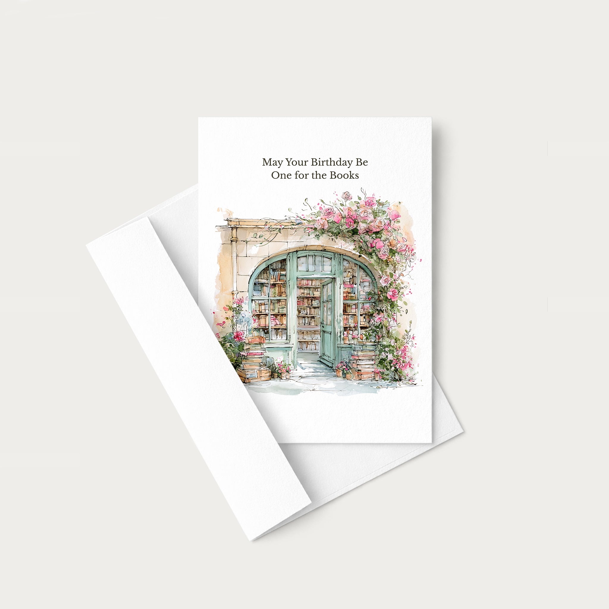 A white greeting card with a watercolor illustration of a bookshop entrance under pink flowers, with the text 'May Your Birthday Be One for the Books,' shown with a white envelope on a light gray background.