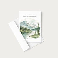 A vertical birthday card with a watercolor mountain lake landscape print, shown with a white envelope on a light warm gray background. The card reads 'Wishing You a Wonderful Birthday' above the scenic illustration.