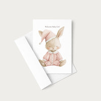 A baby shower card featuring a watercolor illustration of a sleeping bunny in a pink outfit and hat, with the text 'Welcome Baby Girl' above it. The card is shown with its matching white envelope on a light beige background.