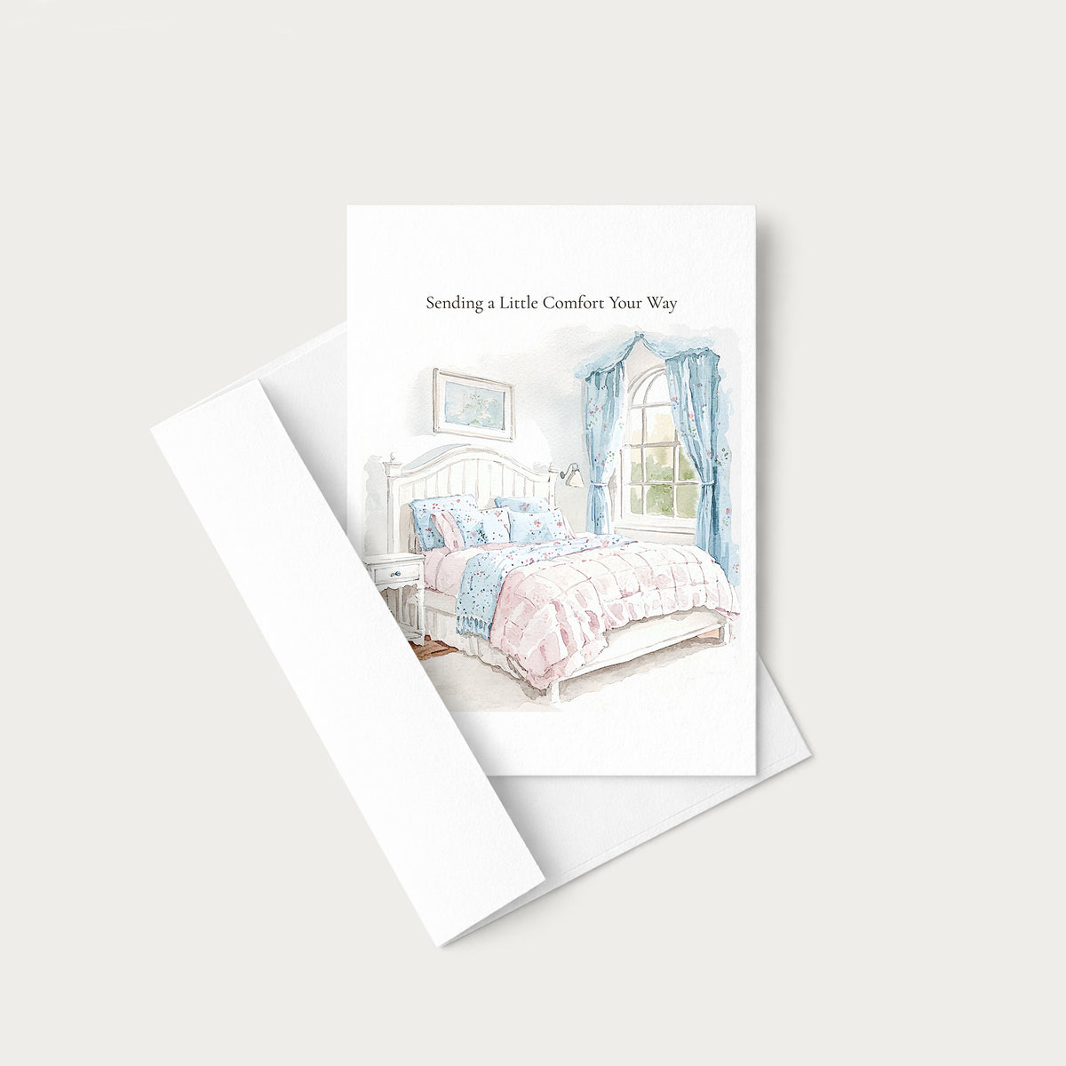A greeting card with a watercolor illustration of a cozy bedroom scene, shown with its white envelope on a light beige background. The card reads 'Sending a Little Comfort Your Way'.
