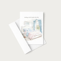 A greeting card with a watercolor illustration of a cozy bedroom scene, shown with its white envelope on a light beige background. The card reads 'Sending a Little Comfort Your Way'.