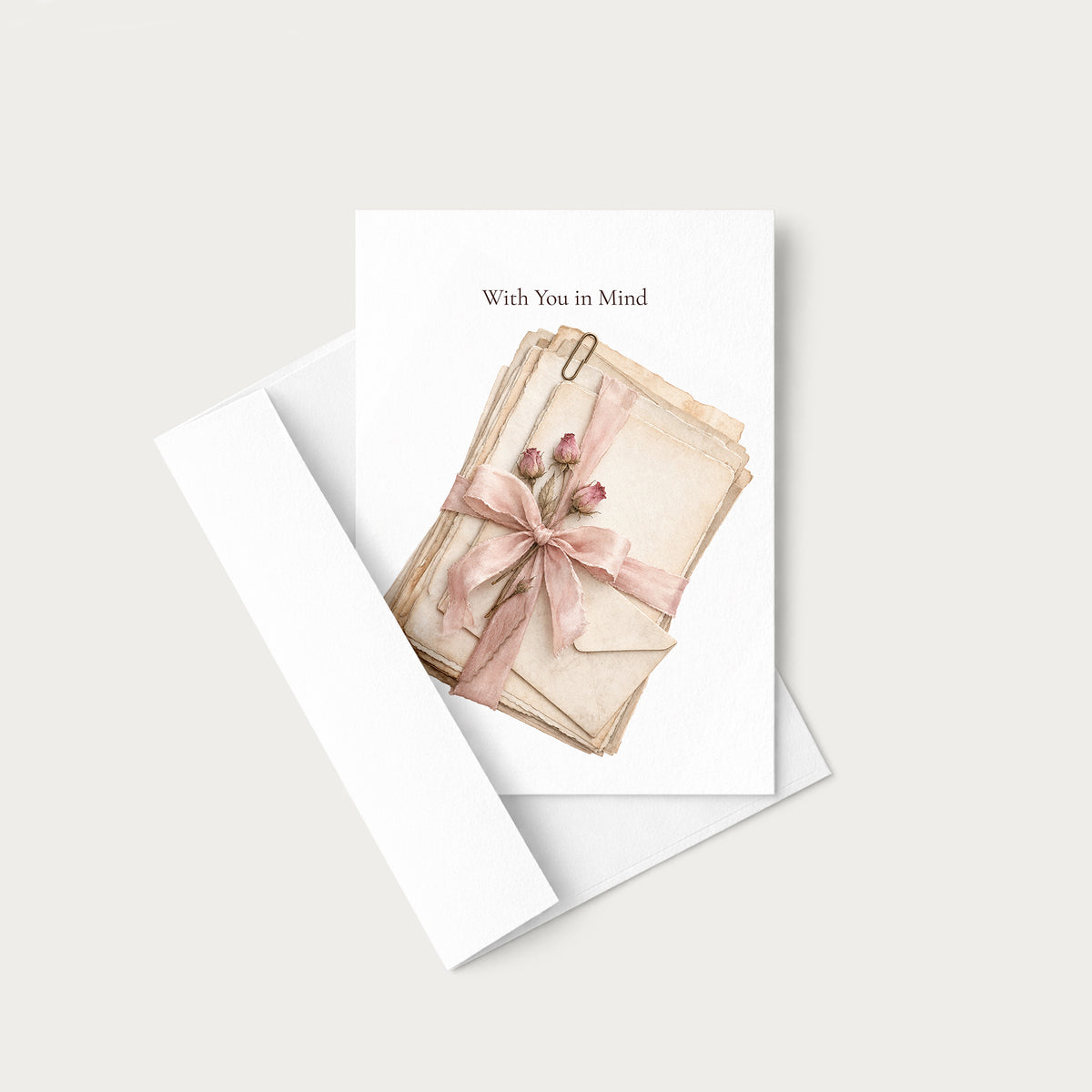 A greeting card with a white envelope, featuring an illustration of stacked vintage paper envelopes tied with a pink ribbon and small flowers, with the text 'With You in Mind' at the top.