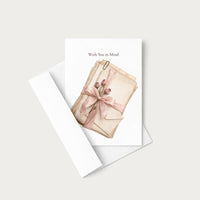 A greeting card with a white envelope, featuring an illustration of stacked vintage paper envelopes tied with a pink ribbon and small flowers, with the text 'With You in Mind' at the top.