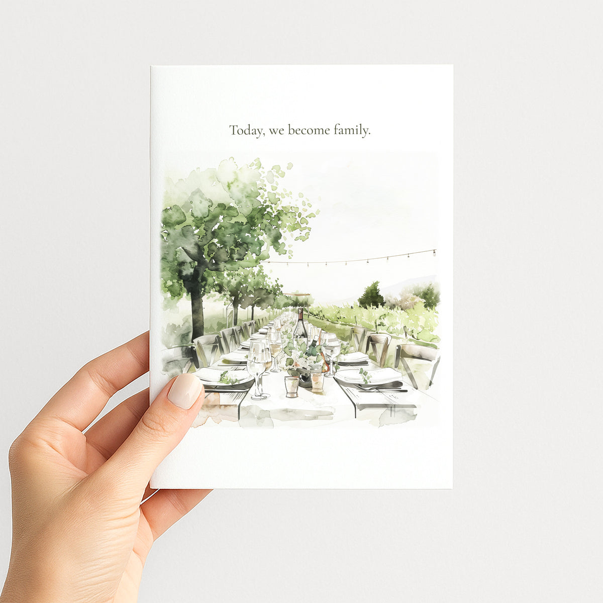 A hand holds a white greeting card featuring a watercolor illustration of an outdoor dining table under trees, with the text 'Today, we become family.' at the top, against a light gray background.