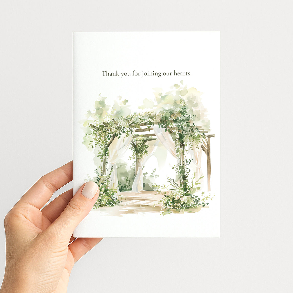 A hand holding a white greeting card with a watercolor illustration of a floral arched pathway and the text 'Thank you for joining our hearts.' on a light gray background.