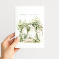 A hand holding a white greeting card with a watercolor illustration of a floral arched pathway and the text 'Thank you for joining our hearts.' on a light gray background.