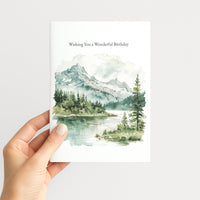 A hand holding a white greeting card with a watercolor landscape of mountains, trees, and a lake. The card reads 'Wishing You a Wonderful Birthday' at the top.