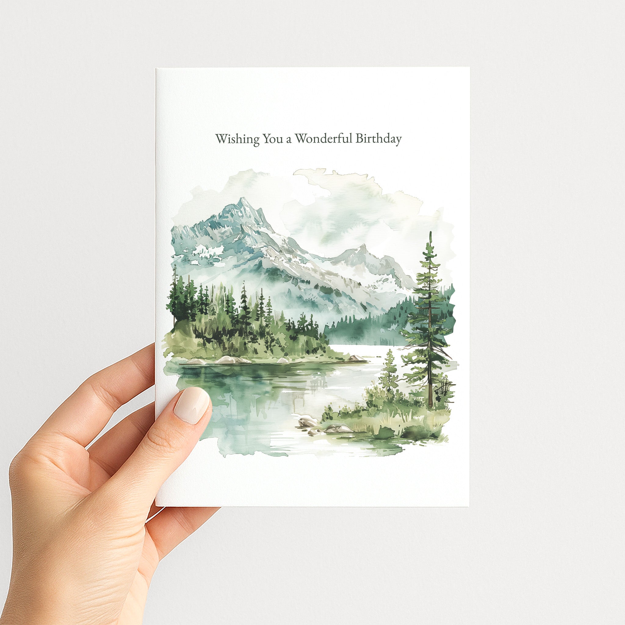 A hand holding a white greeting card with a watercolor landscape of mountains, trees, and a lake. The card reads 'Wishing You a Wonderful Birthday' at the top.