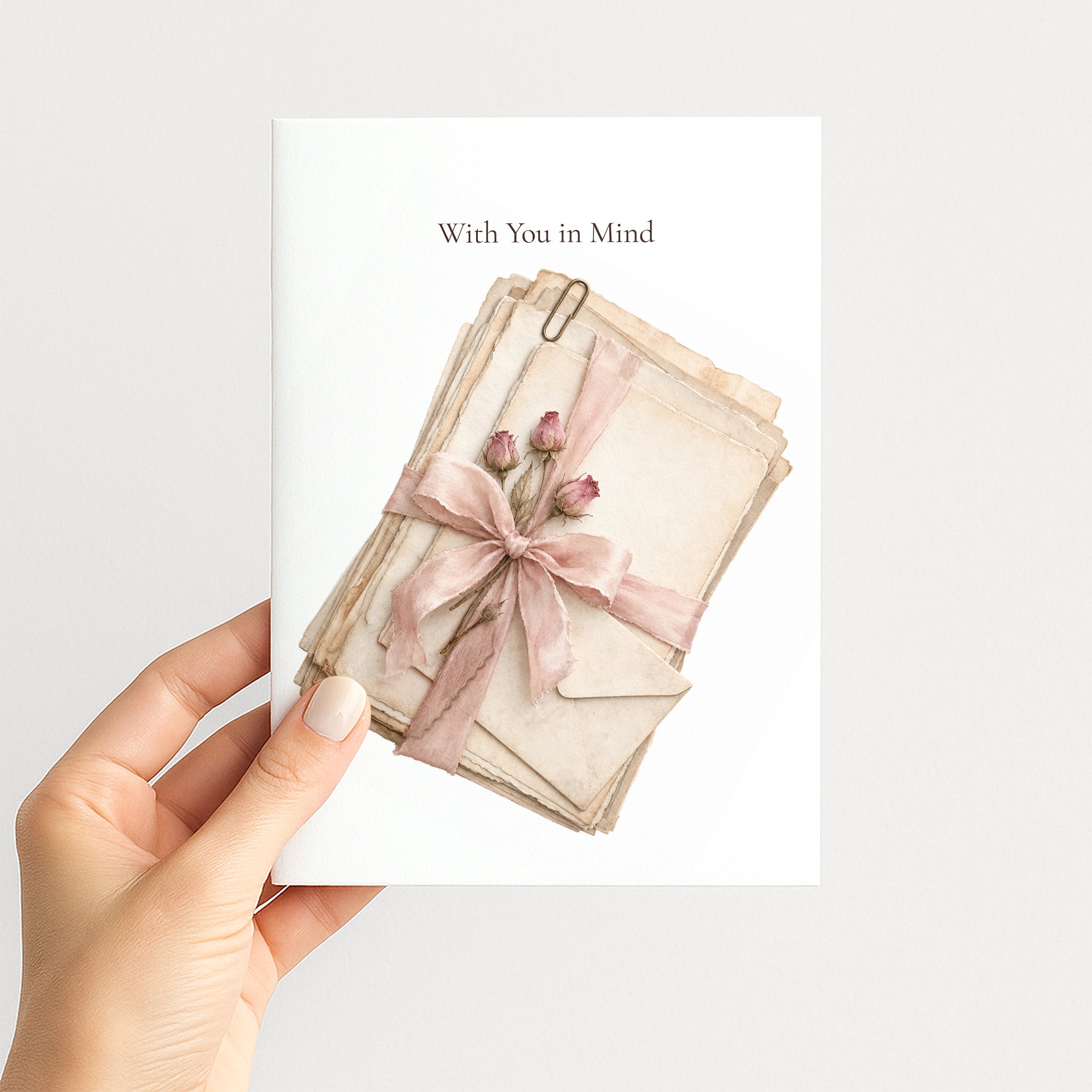 A hand holds a white greeting card against a light gray background. The card has the text 'With You in Mind' above an illustration of stacked beige envelopes tied with a pink ribbon and small pink flower buds.