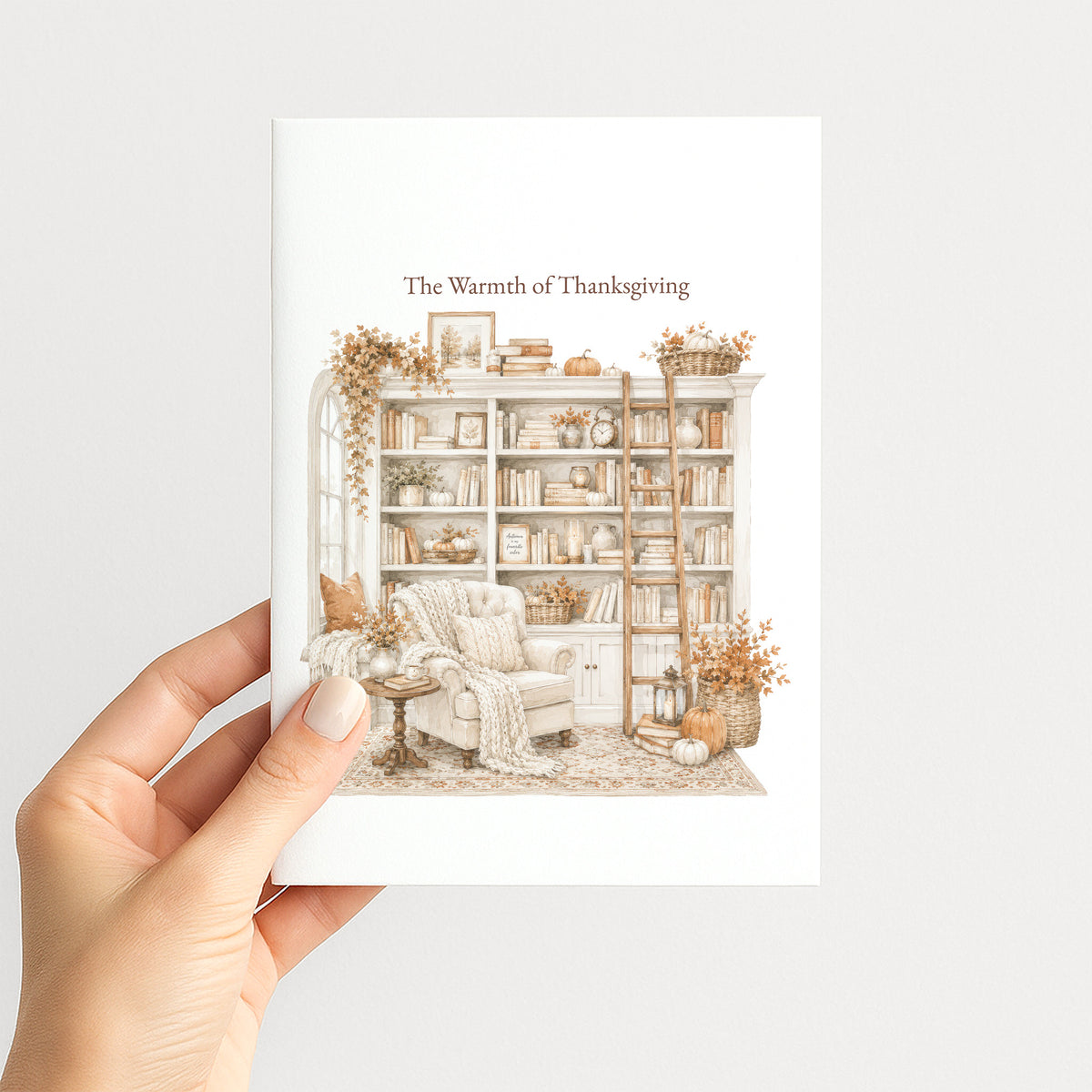 A hand holds a white art print or greeting card featuring a warm, autumnal illustration of a cozy library nook with the text 'The Warmth of Thanksgiving' at the top.