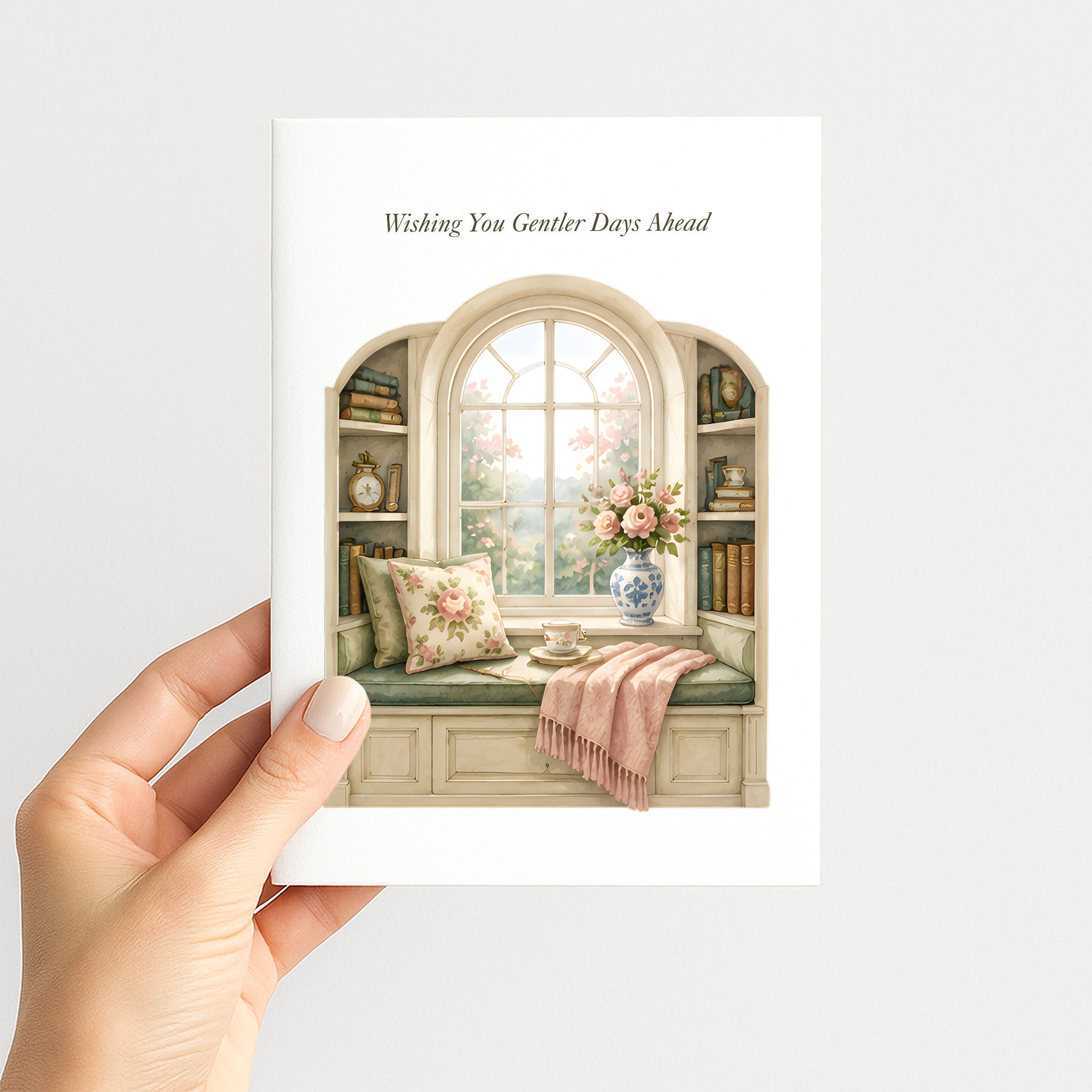A hand holding a greeting card that reads 'Wishing You Greater Days Ahead' with an illustration of a cozy window seat, books, flowers, and a pink throw blanket, shown against a light gray background.