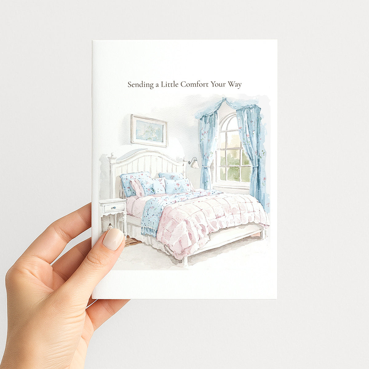 A hand holds a white greeting card featuring a watercolor illustration of a bed with pastel pink and blue bedding, blue curtains, and the text 'Sending a Little Comfort Your Way' at the top.