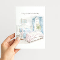 A hand holds a white greeting card featuring a watercolor illustration of a bed with pastel pink and blue bedding, blue curtains, and the text 'Sending a Little Comfort Your Way' at the top.