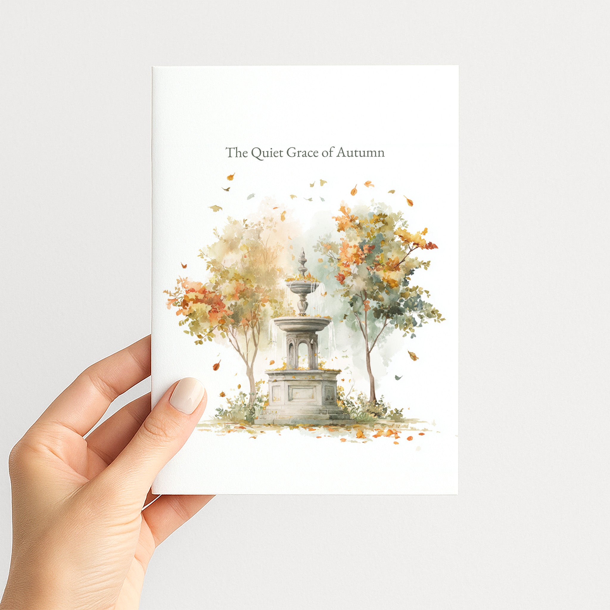 A hand holds a white art print featuring a watercolor illustration of a stone fountain surrounded by autumn trees with orange and yellow leaves. The text at the top reads 'The Quiet Grace of Autumn'.