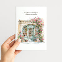 A hand holding a white greeting card with the text 'May Your Birthday Be One for the Books' above a watercolor illustration of a bookshop doorway framed by pink flowers, against a light gray background.