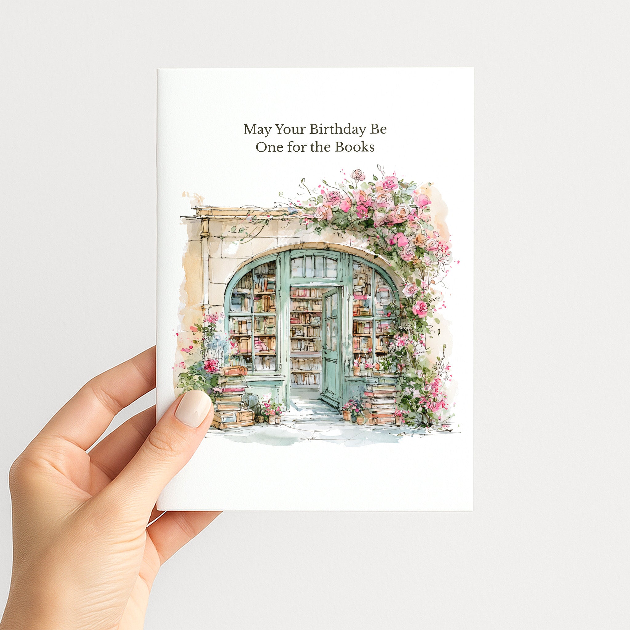 A hand holding a white greeting card with the text 'May Your Birthday Be One for the Books' above a watercolor illustration of a bookshop doorway framed by pink flowers, against a light gray background.