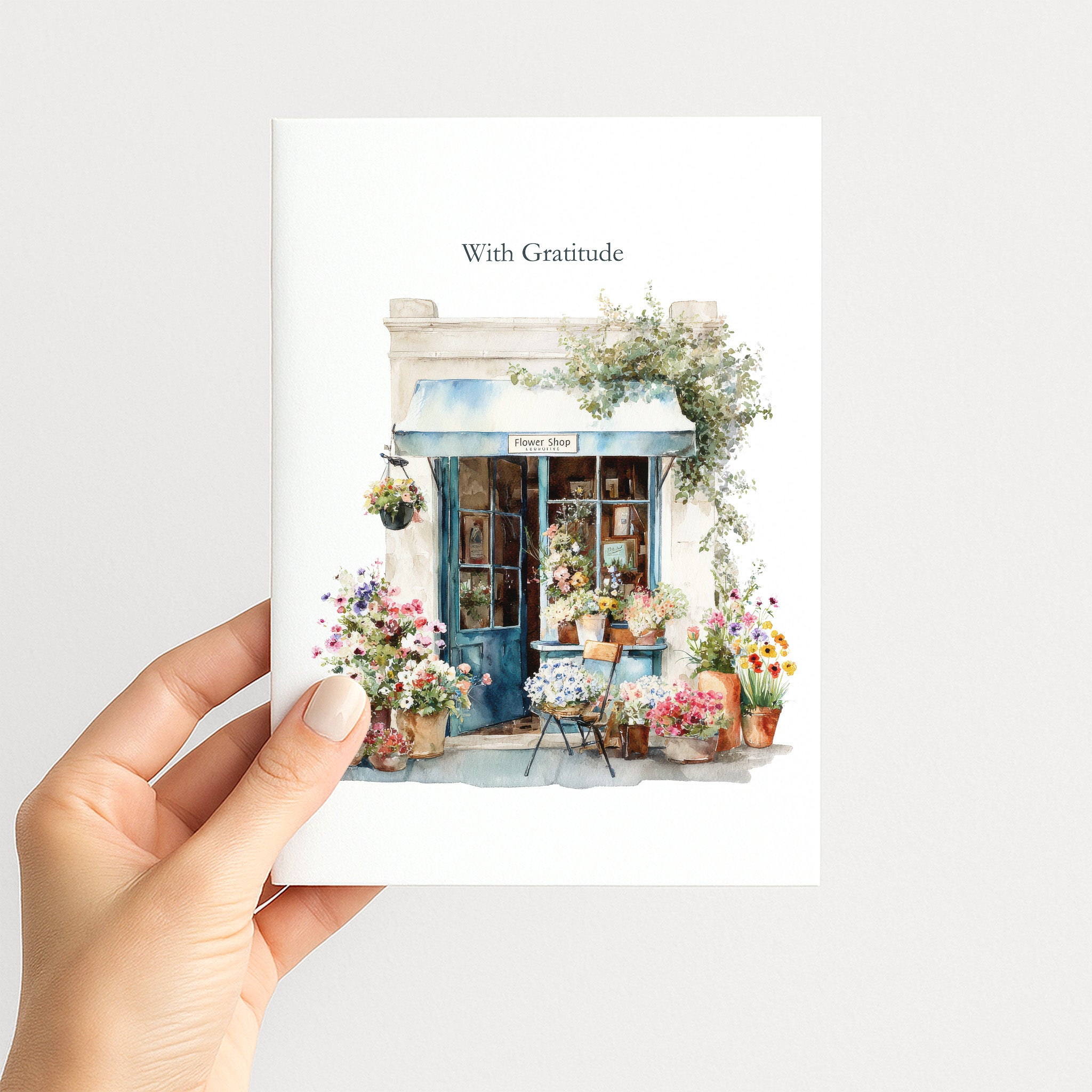 A hand holds a white art print featuring a watercolor illustration of a quaint flower shop with the text 'With Gratitude' above it. The print showcases a blue awning, colorful flowers, and potted plants, set against a clean light gray background.