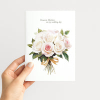 A hand holding a white greeting card with a watercolor bouquet of white and pale pink roses and green leaves, with the printed text 'Dearest Mother, on my wedding day' at the top, against a light gray background.