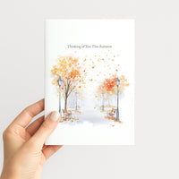 A hand holding a white greeting card featuring a watercolor autumn scene with orange and yellow trees, benches, and falling leaves, with the text 'Thinking of You This Autumn' at the top.