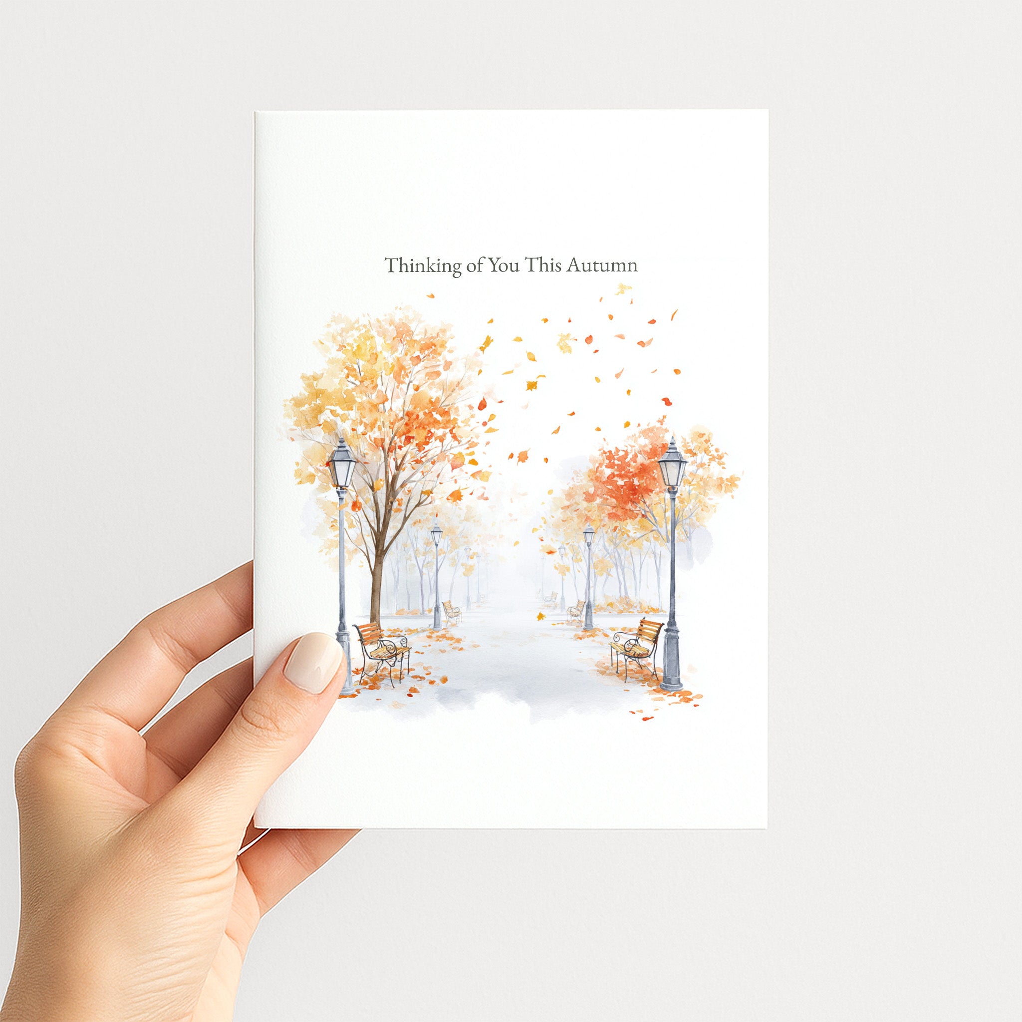 A hand holding a white greeting card featuring a watercolor autumn scene with orange and yellow trees, benches, and falling leaves, with the text 'Thinking of You This Autumn' at the top.