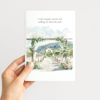 A hand holds a white greeting card featuring a watercolor wedding aisle scene with mountains, floral arches, and the printed quote, 'I can't imagine anyone else walking me down the aisle.'.