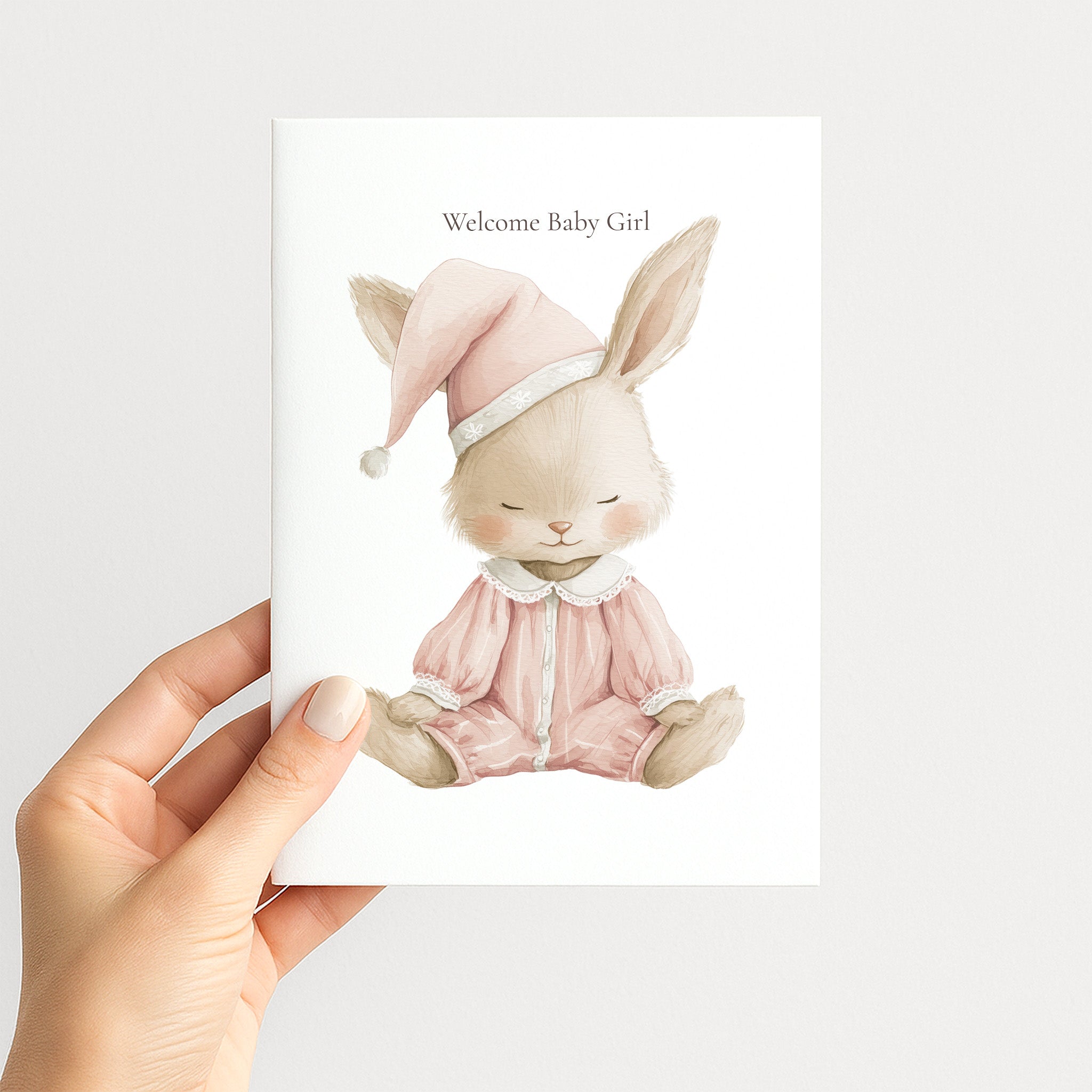 A hand holds a white greeting card featuring a watercolor illustration of a sleeping bunny in pink pajamas and a matching hat, with the text 'Welcome Baby Girl' above it, against a light gray background.