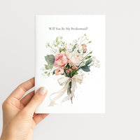 A hand holding a white card that reads 'Will You Be My Bridesmaid?' above a watercolor bouquet of pink and cream flowers with green leaves, against a light gray background.