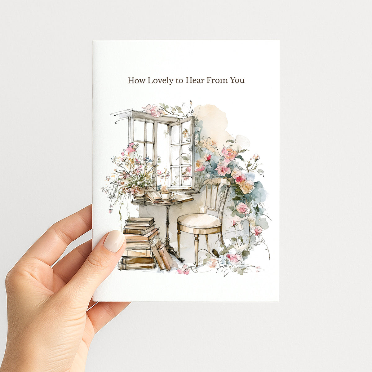 A hand holding a white greeting card with the printed text 'How Lovely to Hear From You' and a watercolor illustration of a cozy window seat with flowers, books, and a chair, against a light gray background.