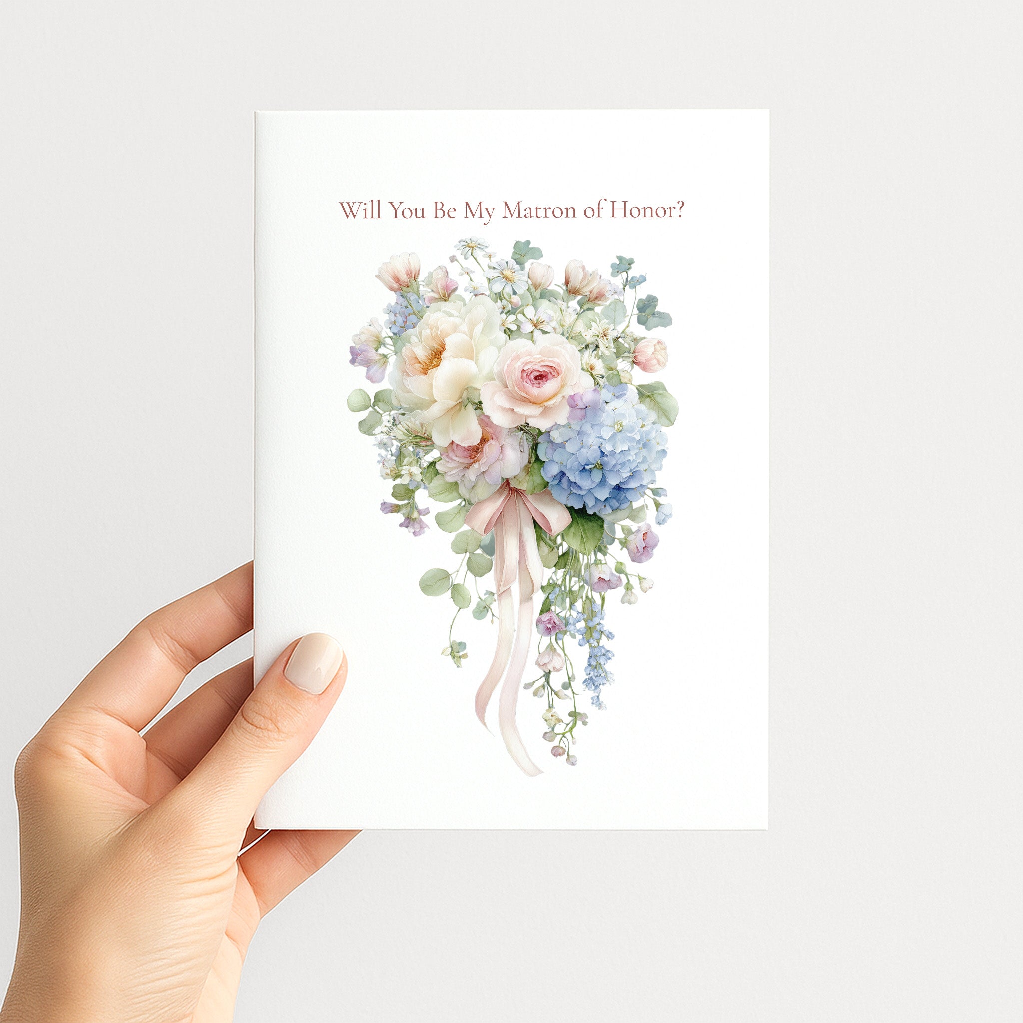 A hand holding a white greeting card that reads 'Will You Be My Matron of Honor?' above a watercolor bouquet of pastel pink, blue, and cream flowers with green leaves, against a light gray background.