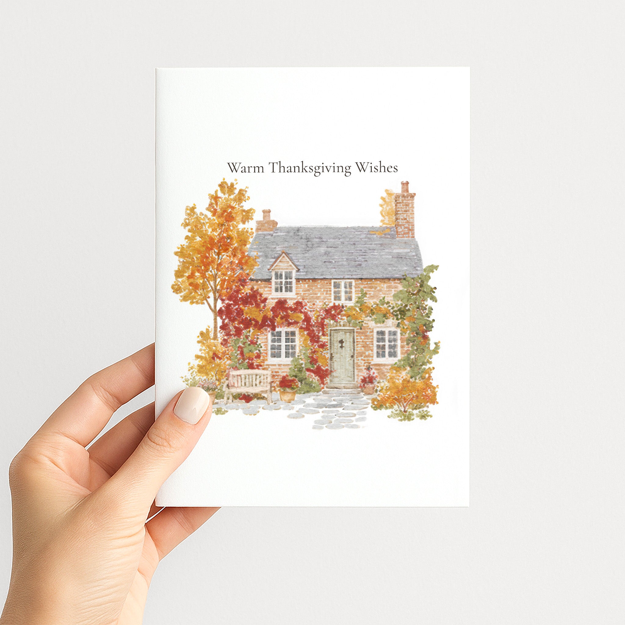 A hand holding a white greeting card with a watercolor illustration of a cottage surrounded by autumn foliage and the text 'Warm Thanksgiving Wishes' at the top, against a light gray background.