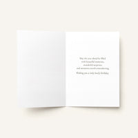 An open white greeting card on a light beige background, with a short printed birthday message in dark gray text on the right panel.