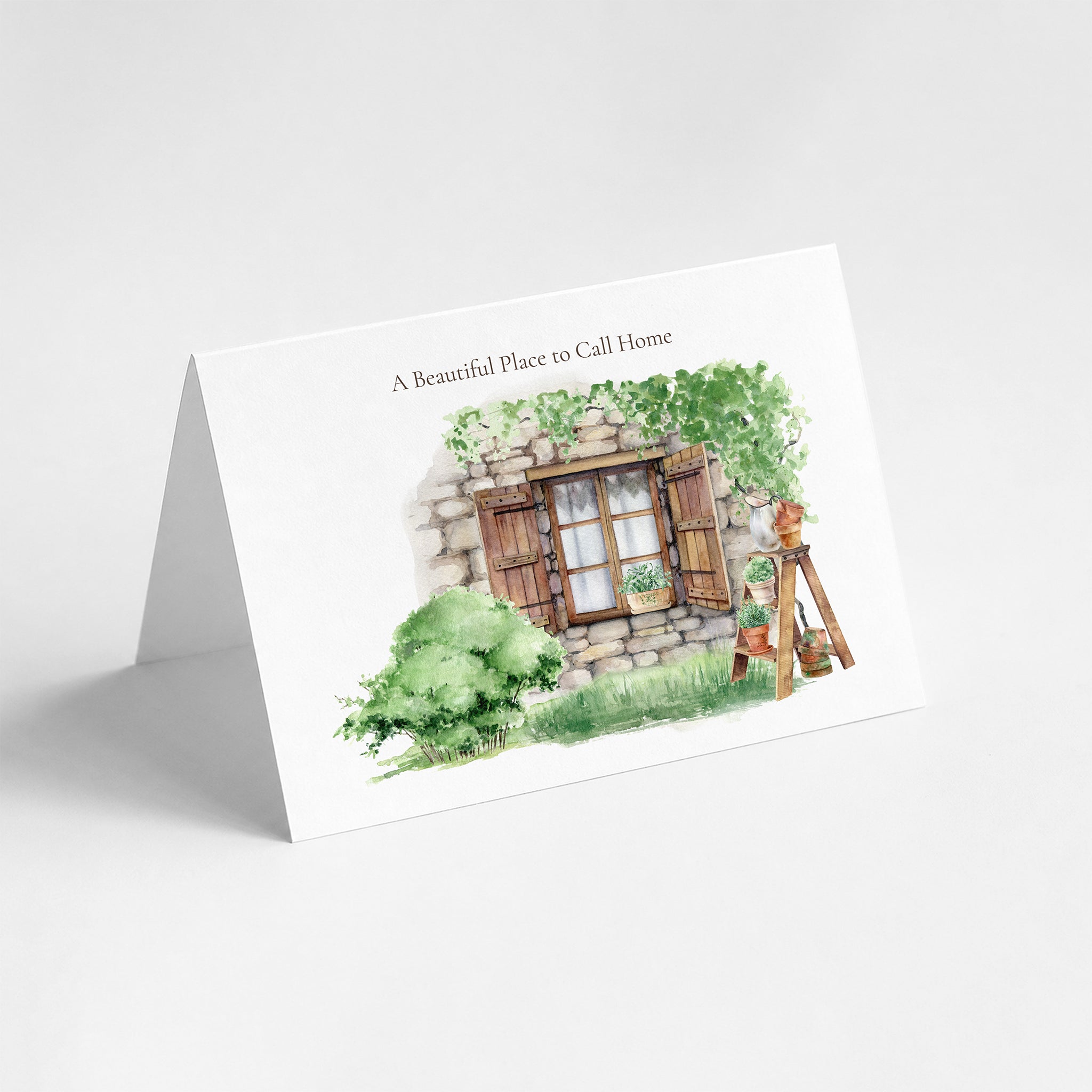 A white A5 landscape greeting card with a watercolor illustration of a stone cottage window, green bushes, and a painter's easel. The card is displayed standing upright on a light gray background, with the text 'A Beautiful Place to Call Home' printed at the top.
