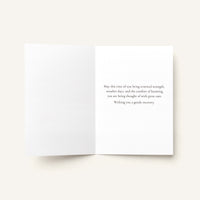 An open white greeting card with a printed birthday message in dark gray text on the right panel, photographed on a light warm beige background.