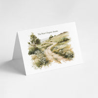 A folded white greeting card standing open on a light gray background, printed with a watercolor illustration of a winding path through grassy hills and the text 'The Next Chapter Awaits'.