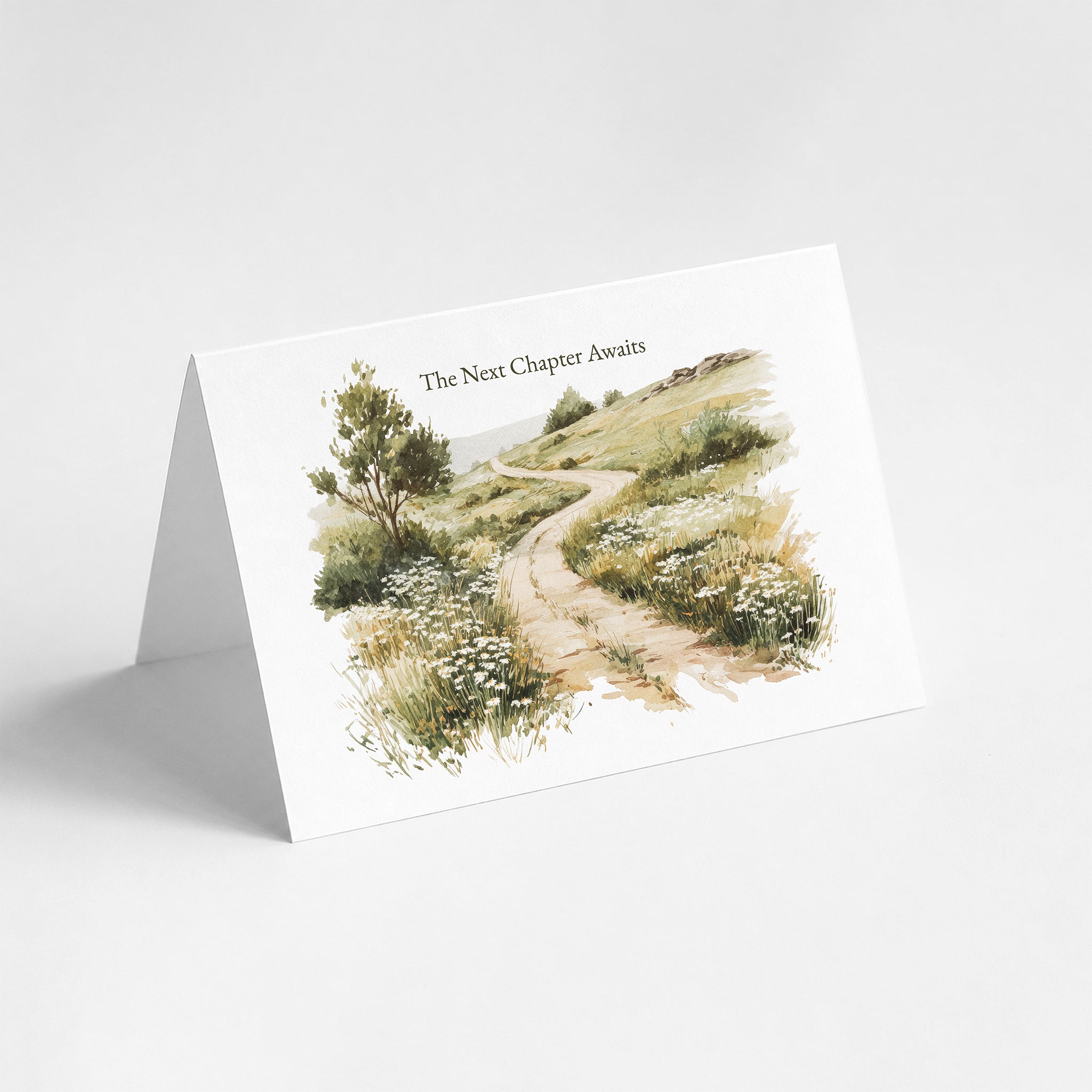 A folded white greeting card standing open on a light gray background, printed with a watercolor illustration of a winding path through grassy hills and the text 'The Next Chapter Awaits'.