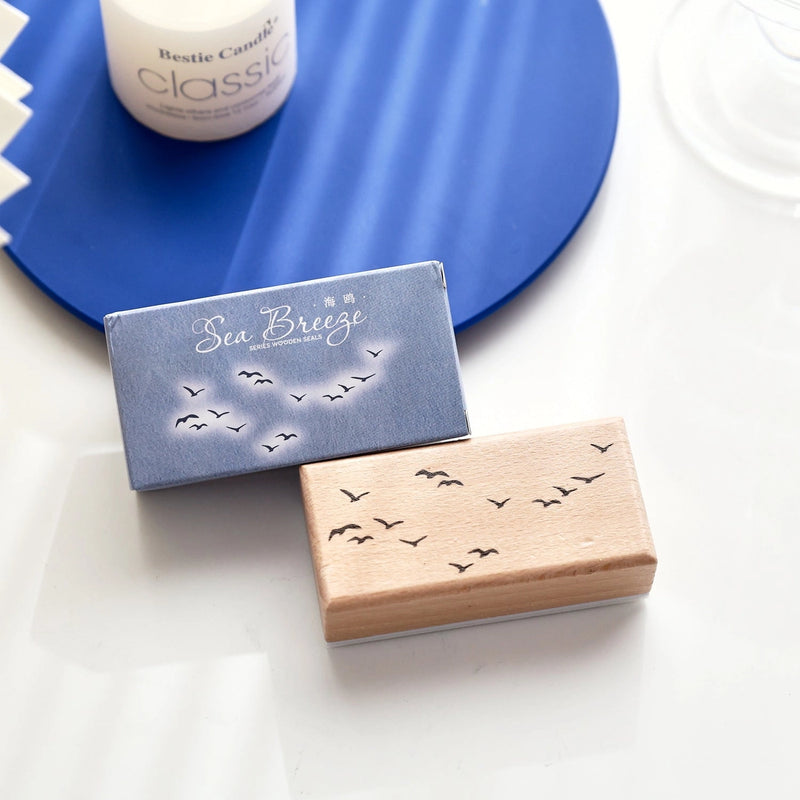 Two rectangular Sea Breeze bath and body products, one in a blue box and one in a light wood box, both printed with small black bird silhouettes, displayed on a white surface with a blue circular prop in the background.