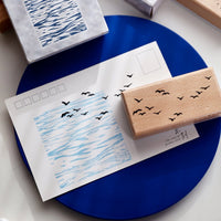 A wooden stamp with black bird silhouettes and a matching blue ink stamp block shown on a white postcard, placed on a blue circular plate.