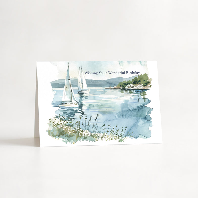 A watercolor-style birthday card standing upright on a white surface against a white background. The card features a scenic coastal painting with sailboats on calm blue water, distant green hills, and the text 'Wishing You a Wonderful Birthday' near the top.