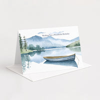 Landscape birthday card with a blue mountain lake and rowboat resting on a white envelope.