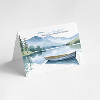 Angled view of a landscape birthday card featuring a peaceful watercolor lake, layered blue mountains, evergreen trees, and a rowboat.
