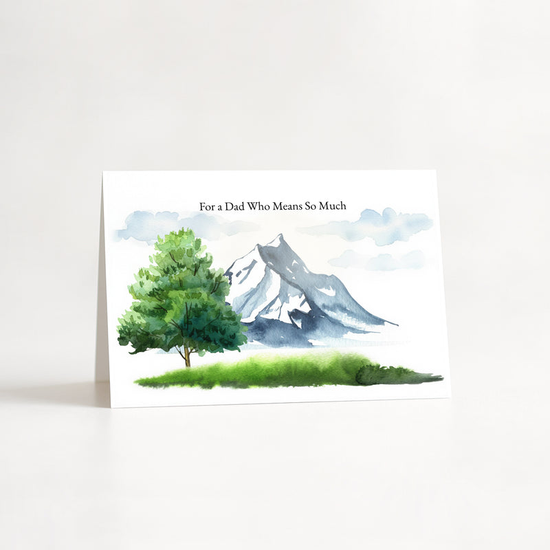A white greeting card standing upright on a white surface, printed with a watercolor landscape of a green tree, grassy foreground, and blue-gray mountain. The text at the top reads, 'For a Dad Who Means So Much'.