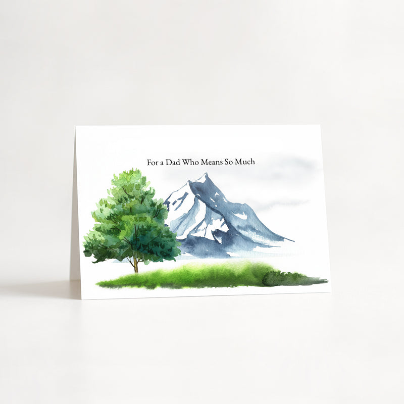 A greeting card standing upright on a white surface, printed with the text 'For a Dad Who Means So Much' above a watercolor-style landscape of a green tree, grass, and blue-gray mountain.