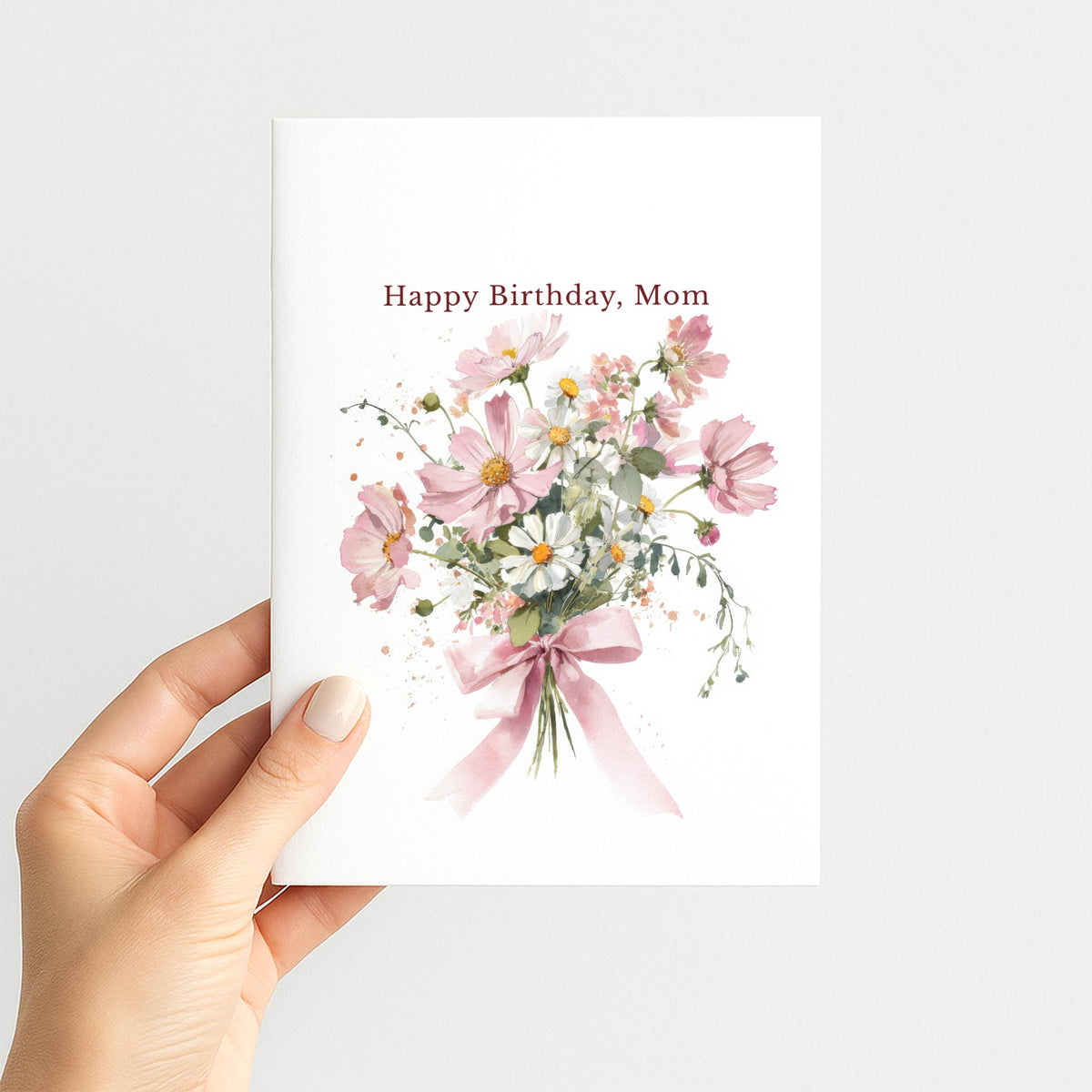 Hand holding a birthday card for Mom featuring a soft pink watercolor bouquet and burgundy lettering.