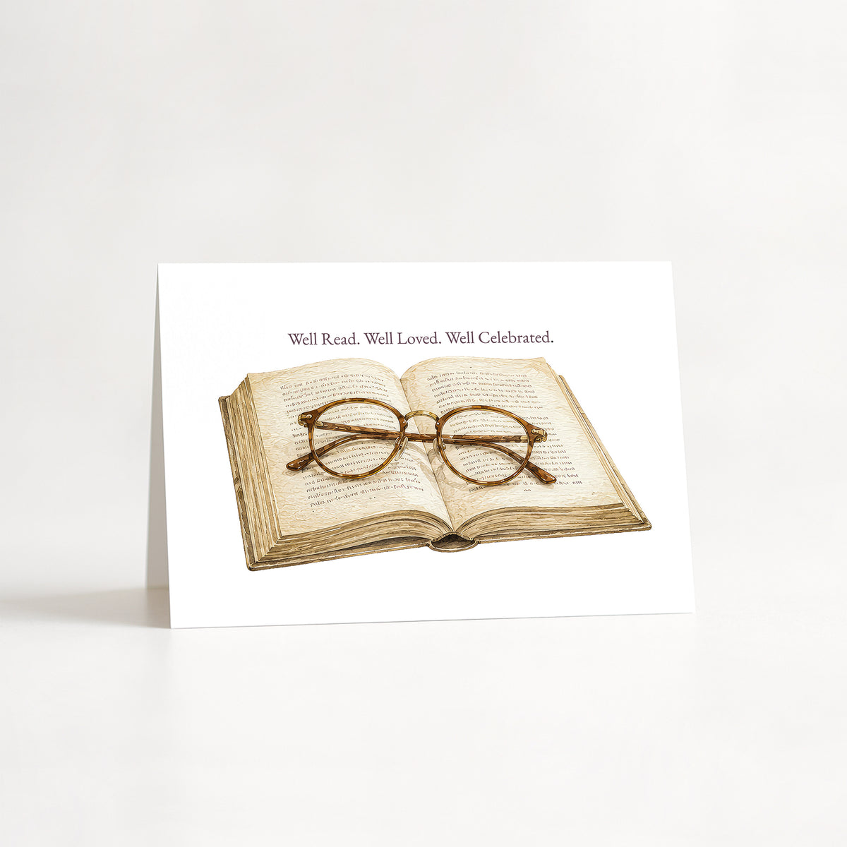 A white greeting card standing upright on a white surface, featuring an illustration of an open book with round wire-frame glasses resting on it, and the text 'Well Read. Well Loved.