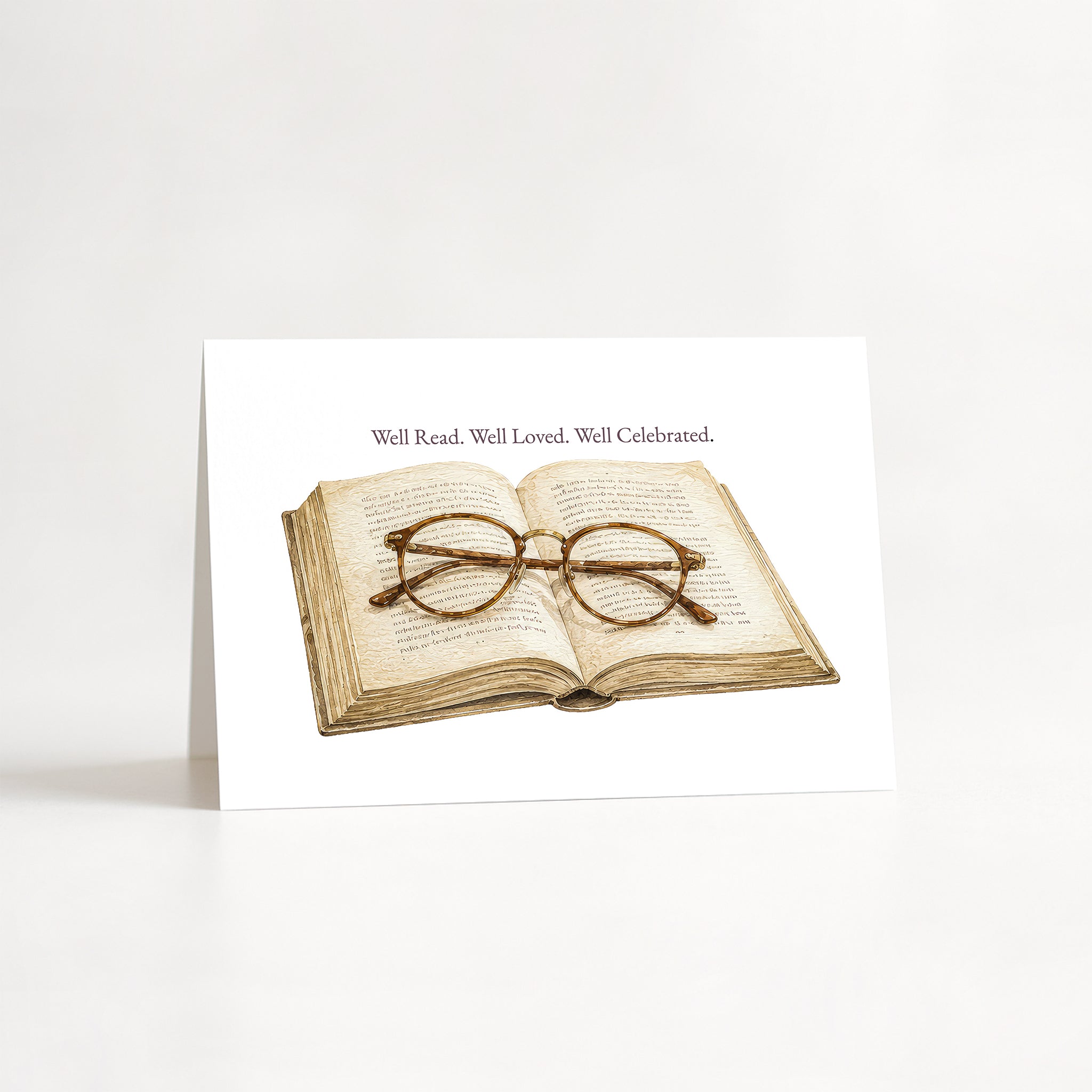 A white greeting card standing upright on a white surface, featuring an illustration of an open book with round wire-frame glasses resting on it, and the text 'Well Read. Well Loved.